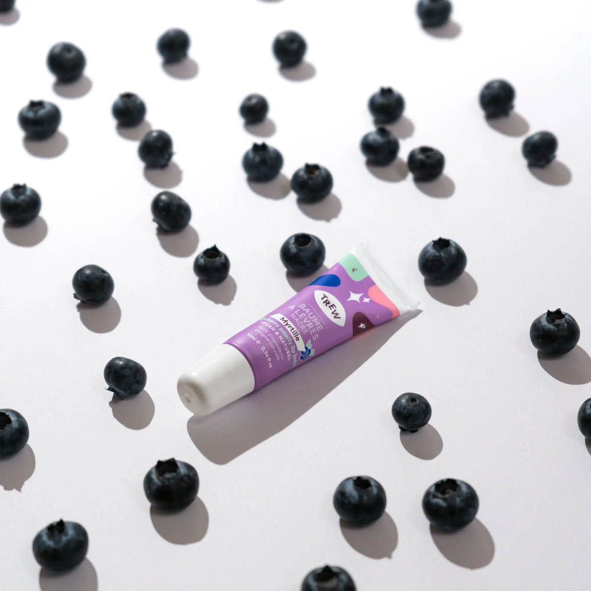 A tube of purple blueberry tinted lip balm centered among scattered fresh blueberries on a white background.