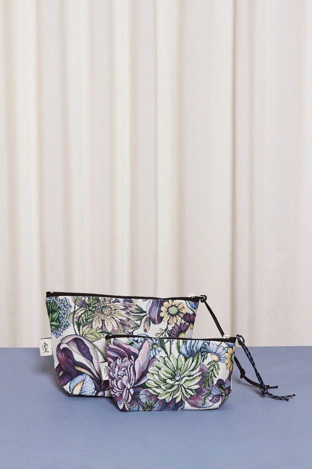 Two zipper pouches in jacquard fabric with a blue chrysanthemum floral pattern, set against a cream curtain backdrop.