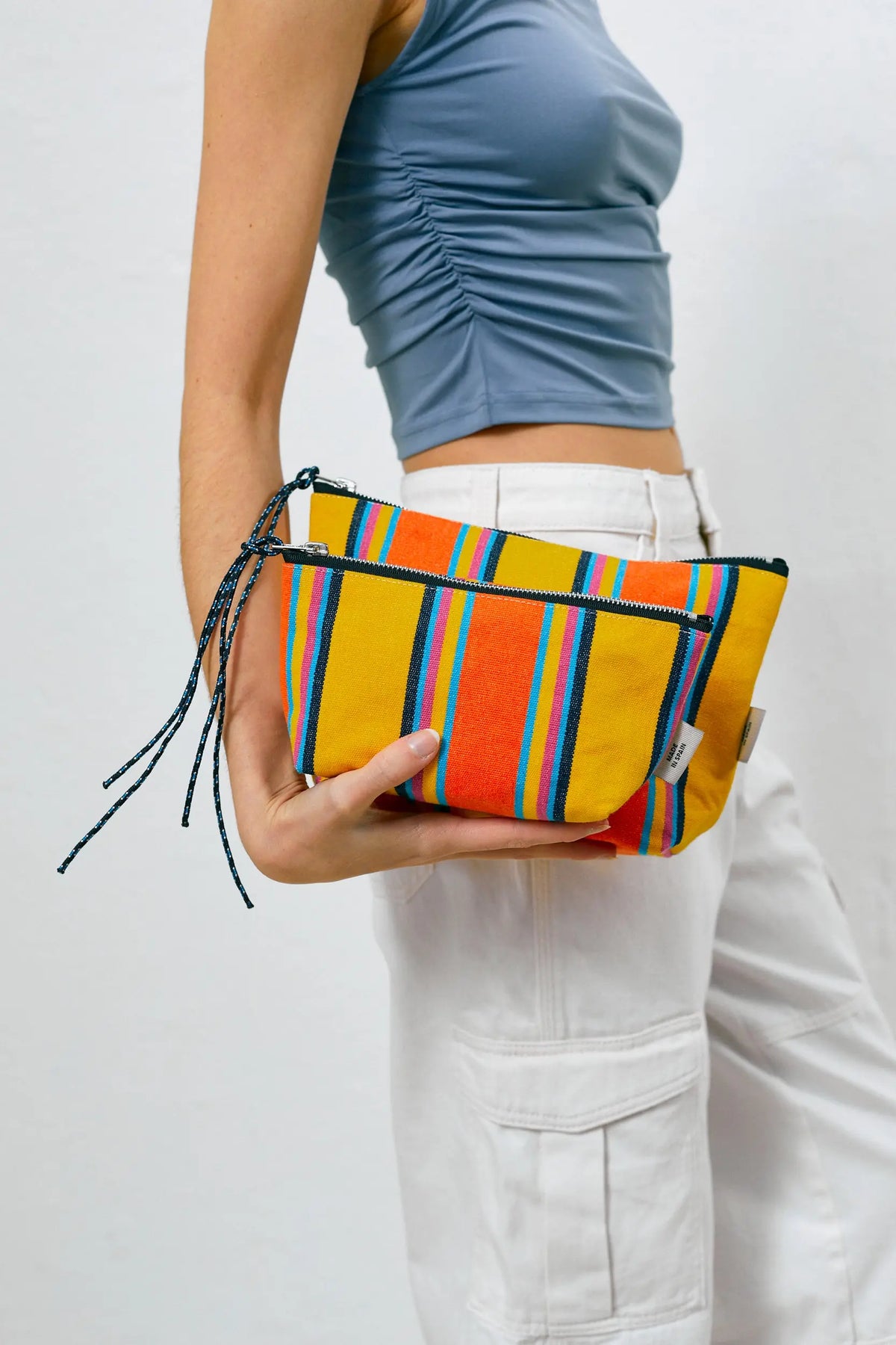 A person holds two yellow, orange, and blue striped canvas pouches with matching rope zipper pulls.