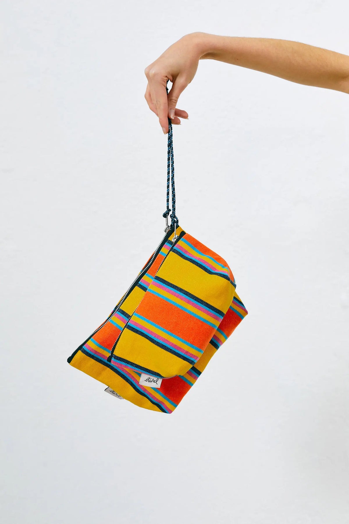 Hand holding a rectangular canvas toiletry bag with vibrant yellow, orange, blue, and pink stripes and a rope handle.