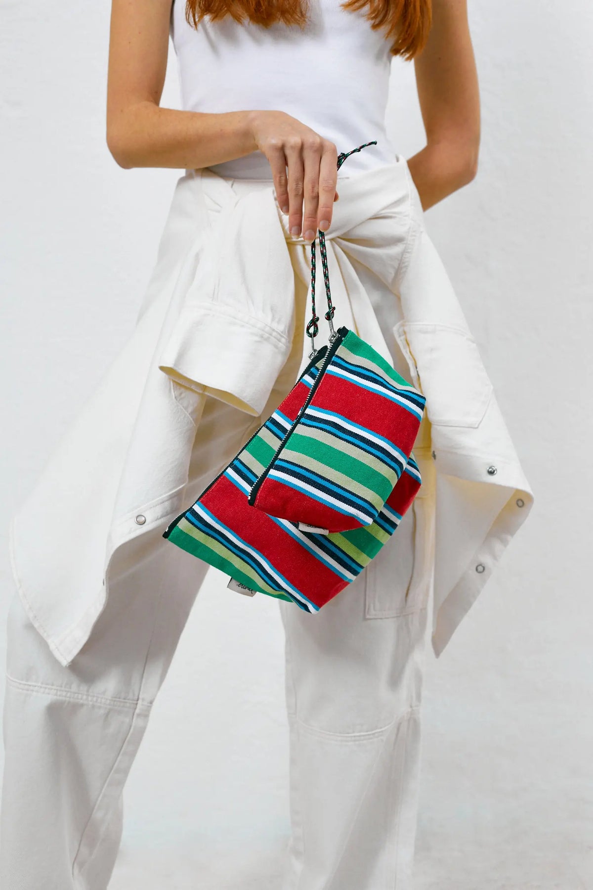 Neceser de lona estilo hamaca con rayas rojas, verdes y azules, con cierre de cremallera y asa de cuerda ajustable.