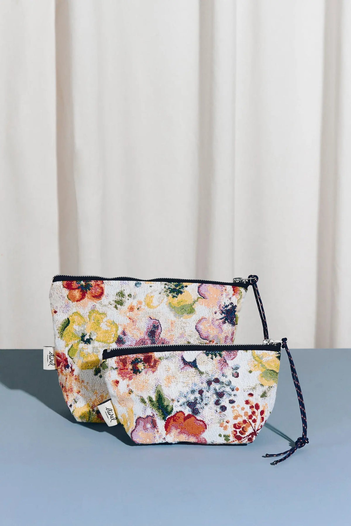 Dos neceseres de tela jacquard con estampado floral multicolor, cierre de cremallera y tirador de cordón azul.