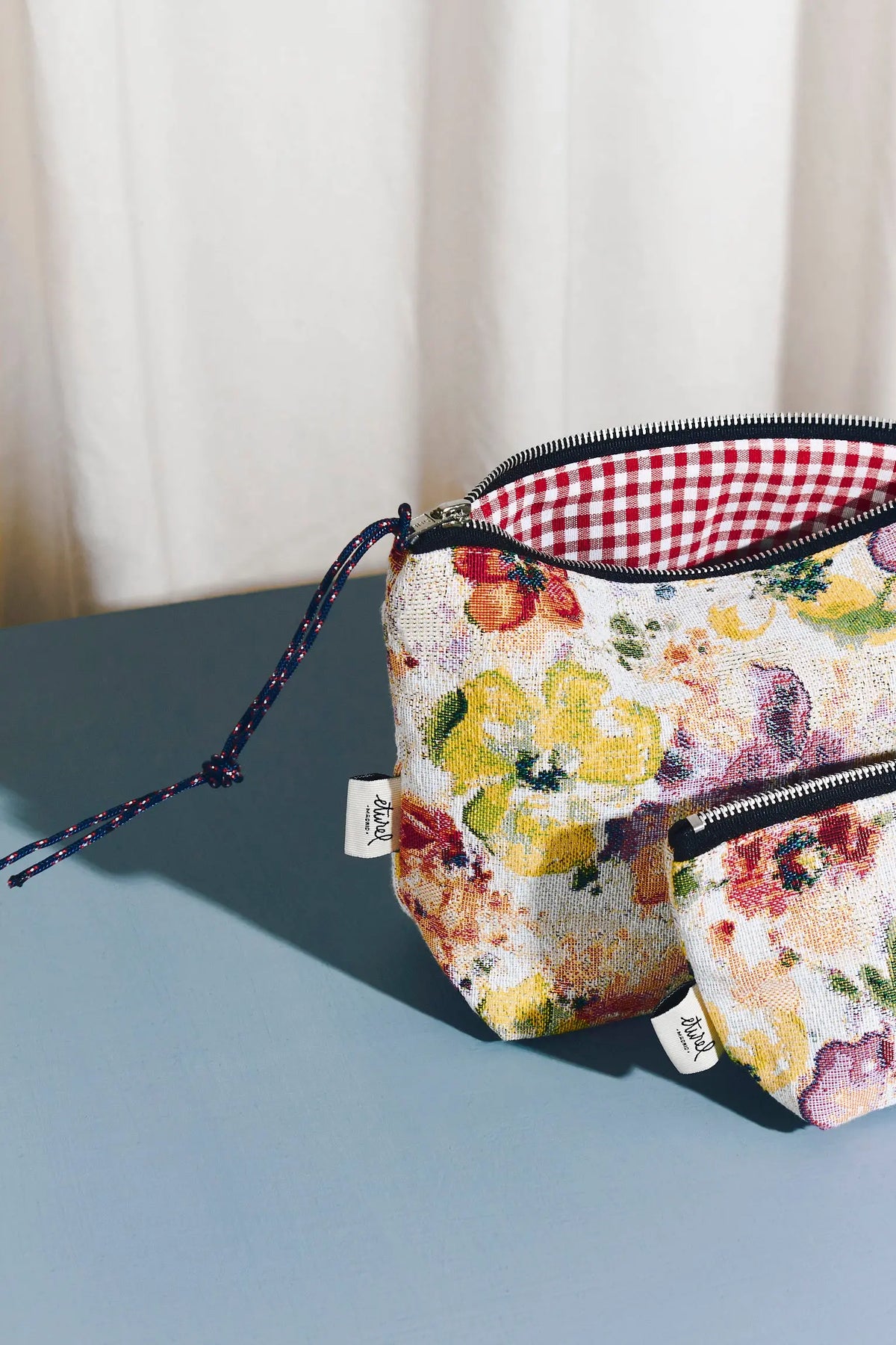 Poppy-patterned jacquard toiletry bags with red gingham lining, zipper closure, and rope pull on a blue surface.