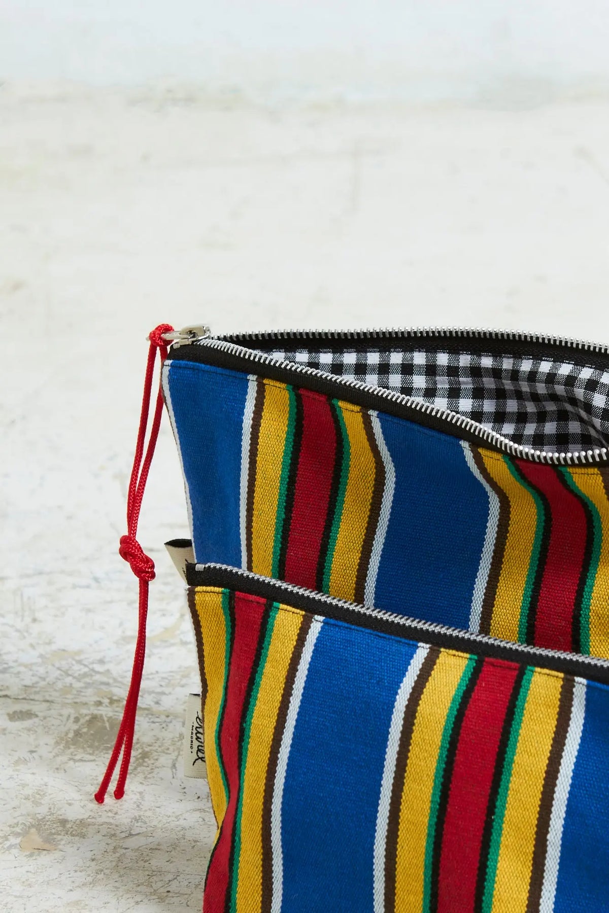 Neceser de lona a rayas azules, amarillas y rojas con cremallera y tirador rojo. Forro interior de cuadros vichy.