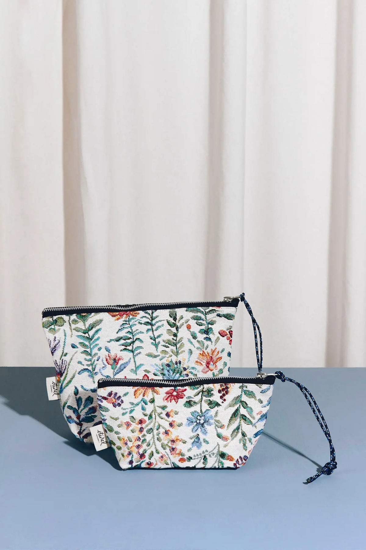 Dos neceseres de tela jacquard con estampado floral de azaleas en tonos vivos, cierre de cremallera y cordón lateral.