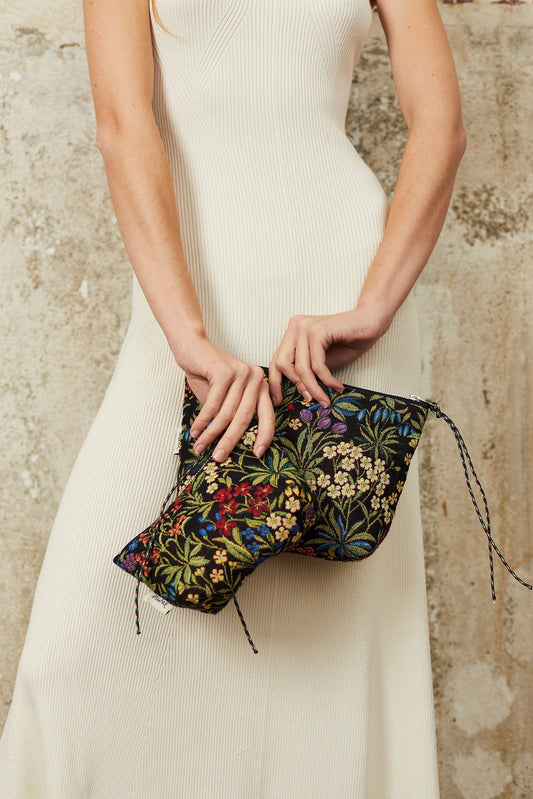 Neceser de tela jacquard con estampado de flores coloridas sobre fondo negro, sostenido por una mujer en vestido blanco.
