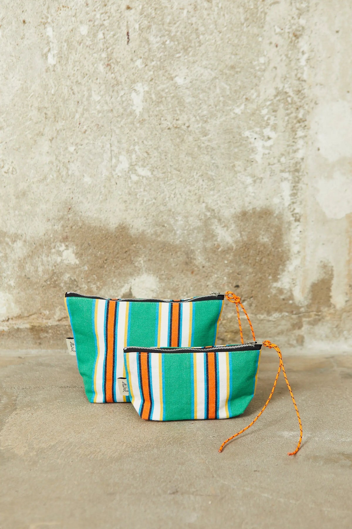 Two green, white, and orange striped canvas pouches with orange cord zipper pulls against a rustic stone wall.