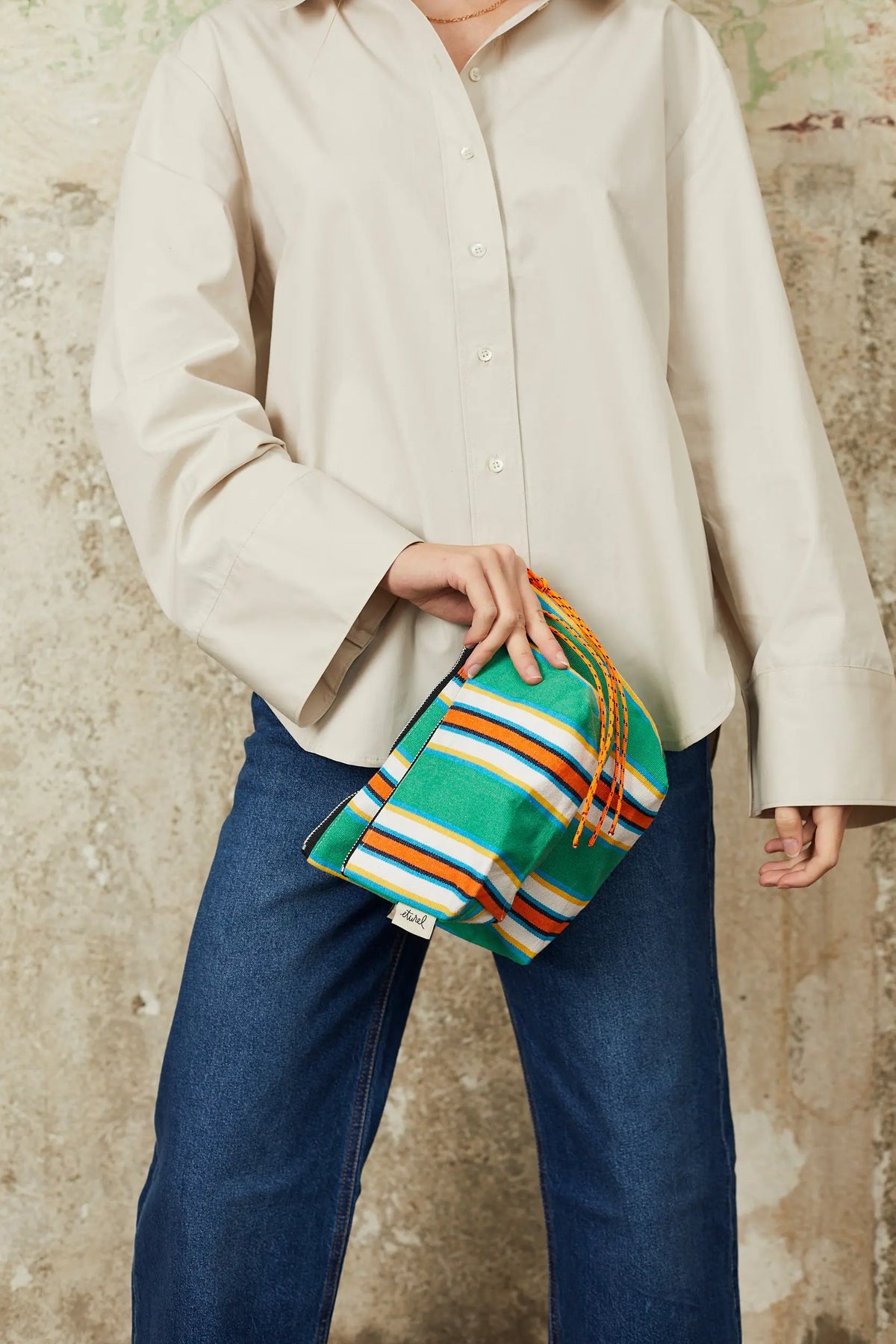 Green striped canvas toiletry bag with orange cord drawstring held by a person in a beige shirt and blue jeans.