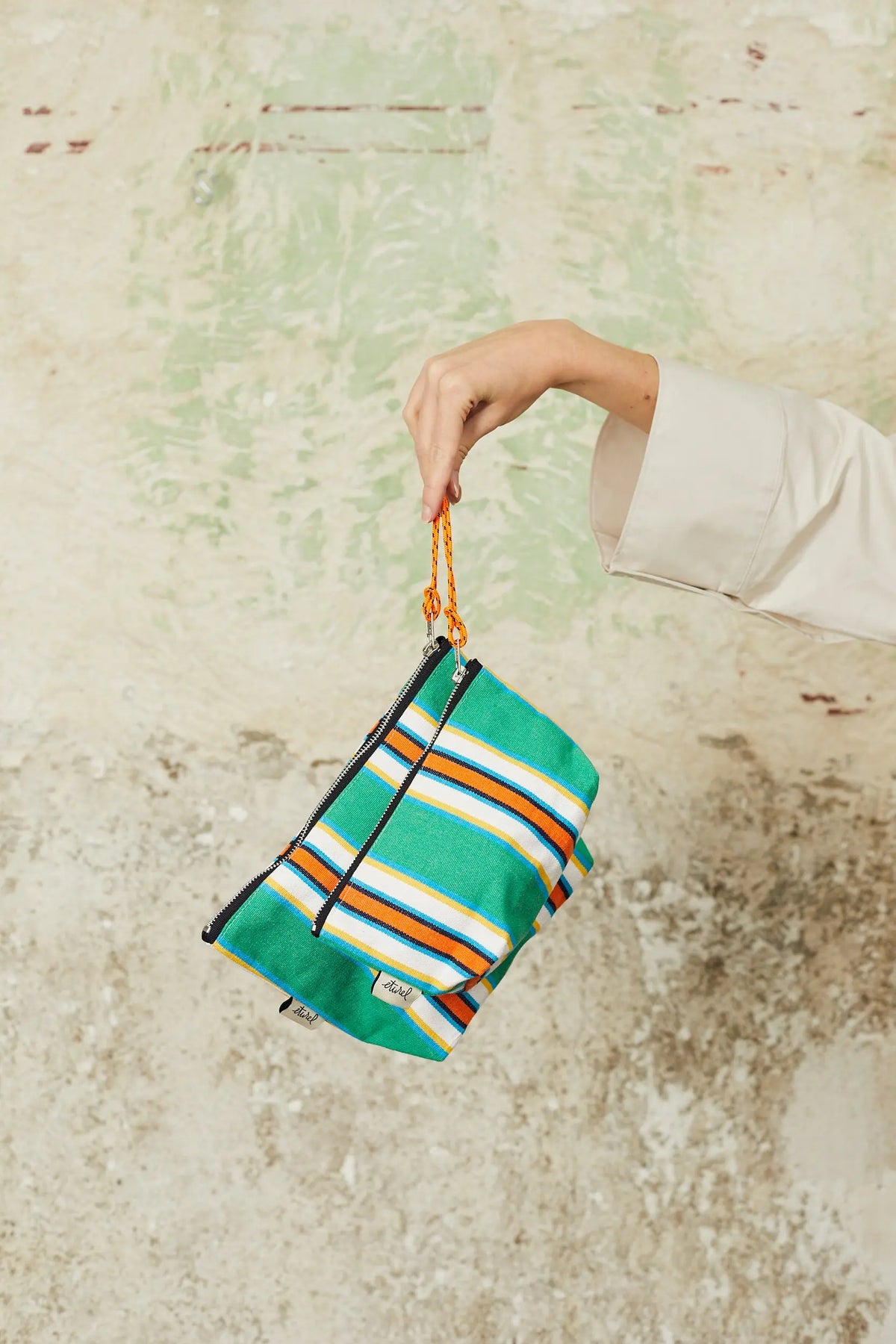 Green and white striped canvas toiletry bag with orange accents, black zipper, and a hanging cord, held by a hand.