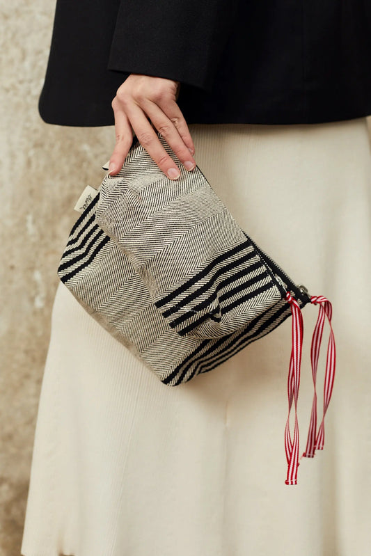 Neceser de sarga con estampado de espiga negro y beige, cierre de cremallera con cintas rojas y blancas, estilo artesano.