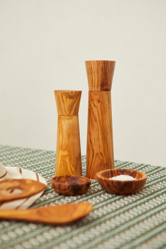 Dos molinillos de madera de olivo con veteado natural sobre mantel estampado, acompañados de cuencos de madera.