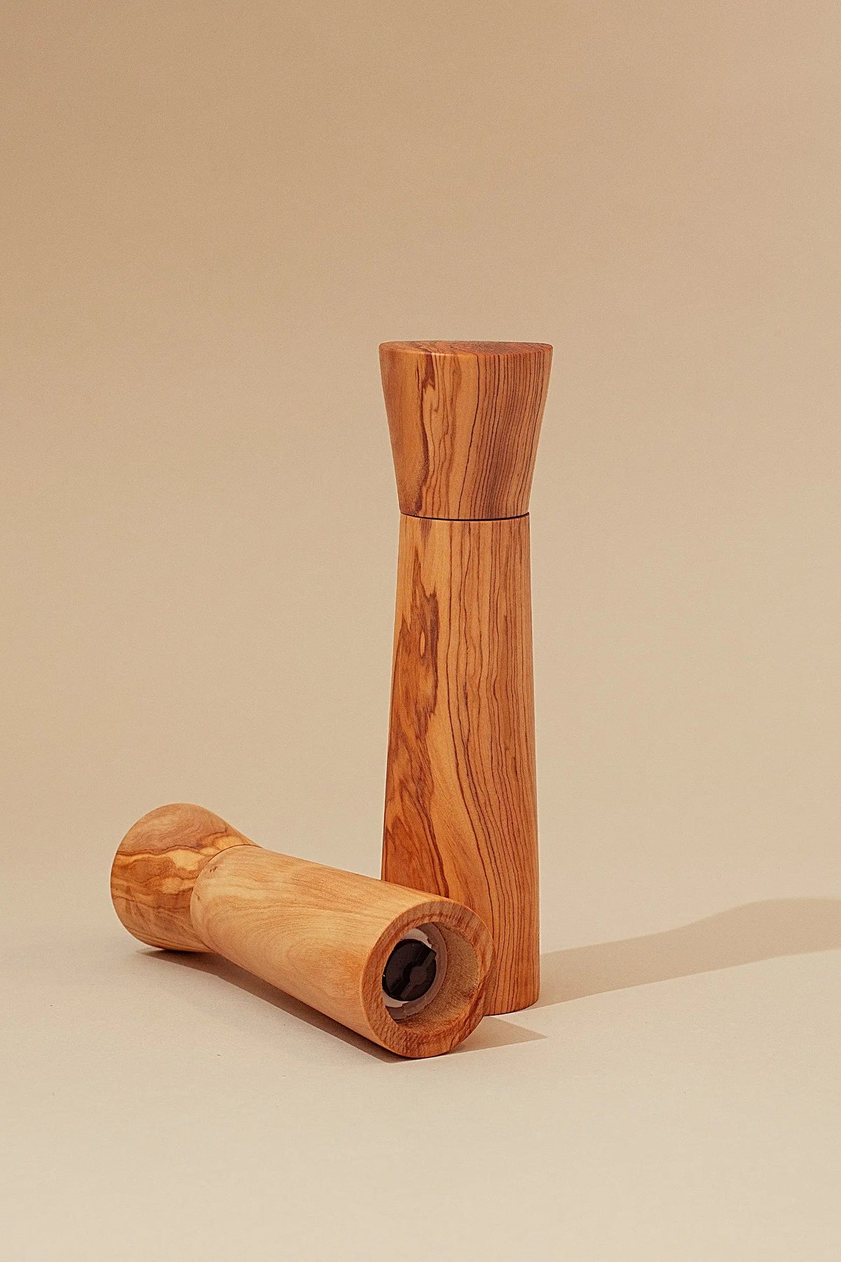 Dos molinillos de sal y pimienta de madera de olivo con veteado natural sobre un fondo beige neutro.