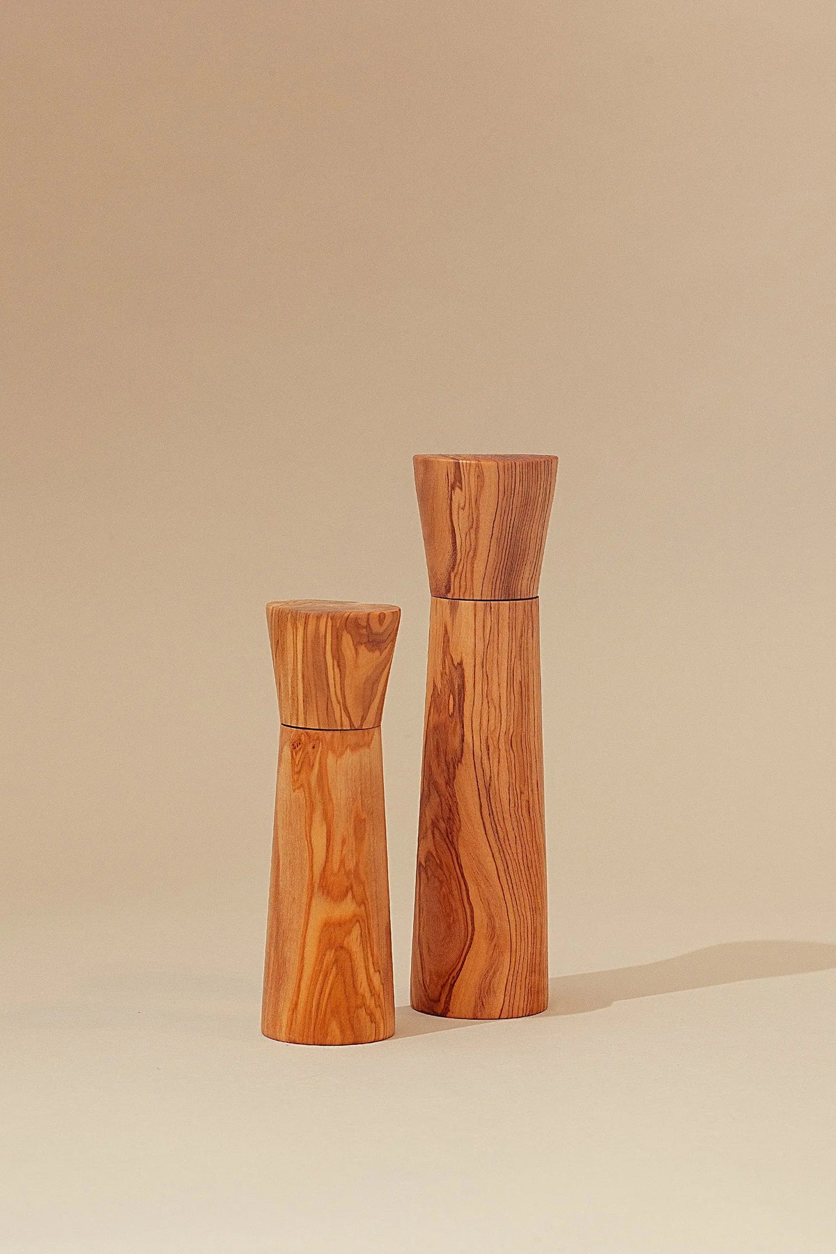 Dos molinillos de madera de olivo con veteado natural y diseño minimalista sobre fondo color arena.