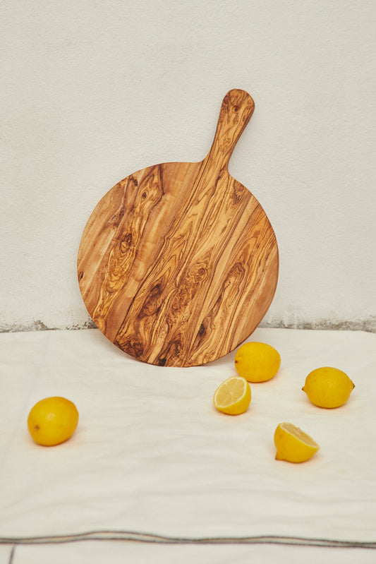 Tabla redonda de madera de olivo con mango, con vetas naturales, apoyada sobre tela clara junto a varios limones.
