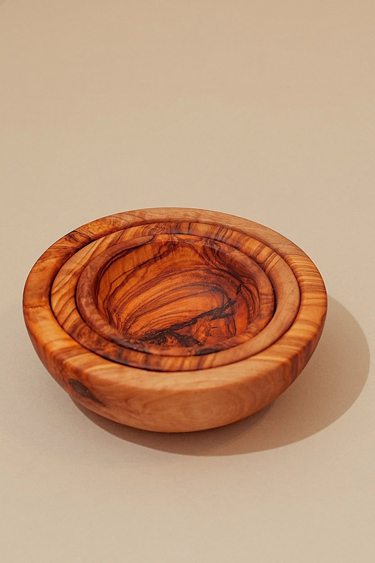 Two nested olive wood bowls with a rich, natural grain and warm brown tones, set against a neutral beige background.