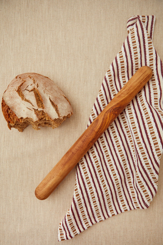 Rodillo de cocina de madera de olivo con veteado natural sobre un paño de rayas junto a una hogaza de pan artesano.
