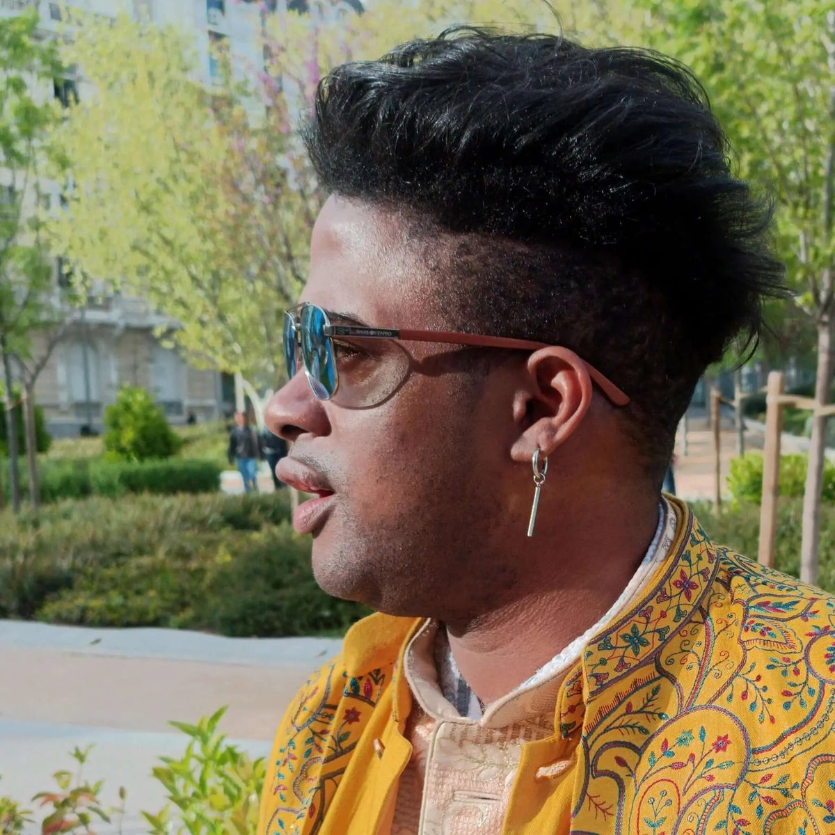 Hombre con gafas de sol de lentes azules y patillas de madera, con chaqueta amarilla estampada en un parque.