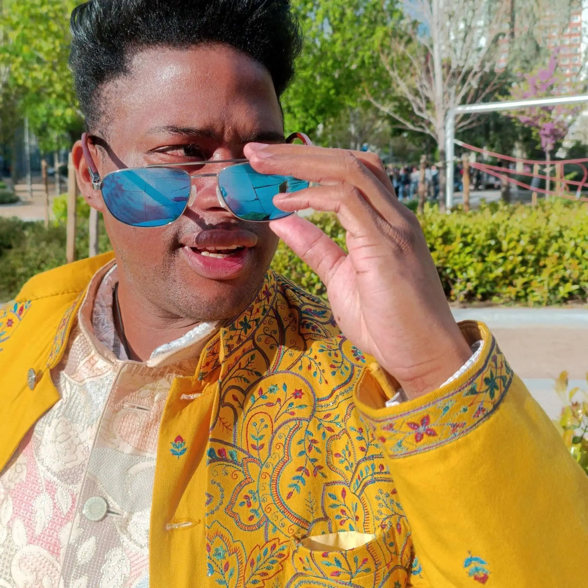 Hombre con chaqueta amarilla y gafas de sol con lentes azules espejadas y patillas de madera en un parque soleado.