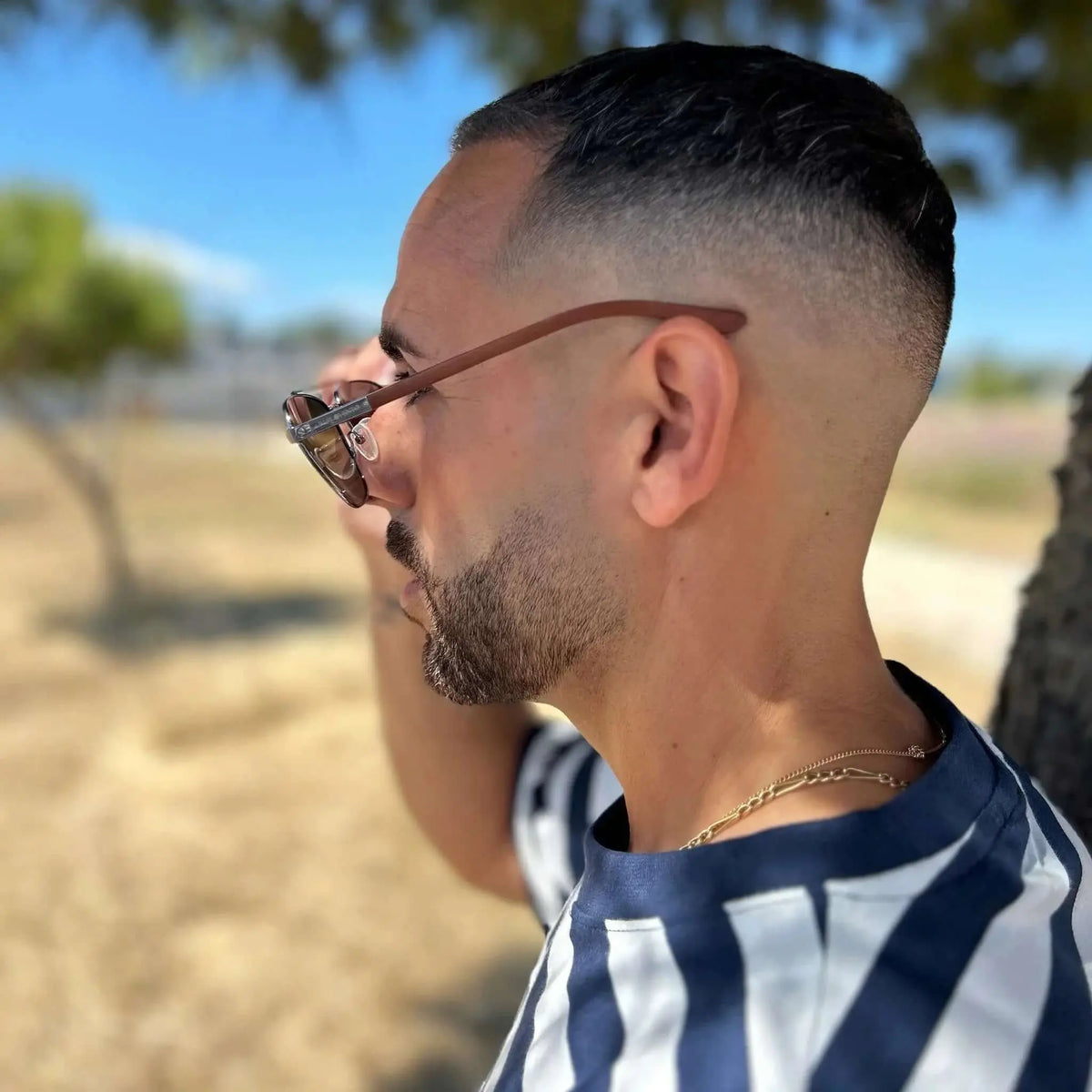 Hombre con gafas de sol de cristales azules y patillas de madera, vistiendo camiseta a rayas en entorno exterior.