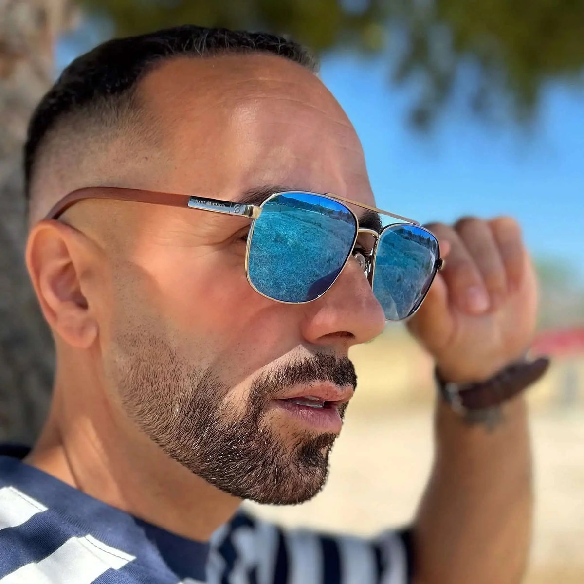 Hombre con gafas de sol aviador, montura metálica, lentes azules espejadas y patillas de madera en entorno soleado.