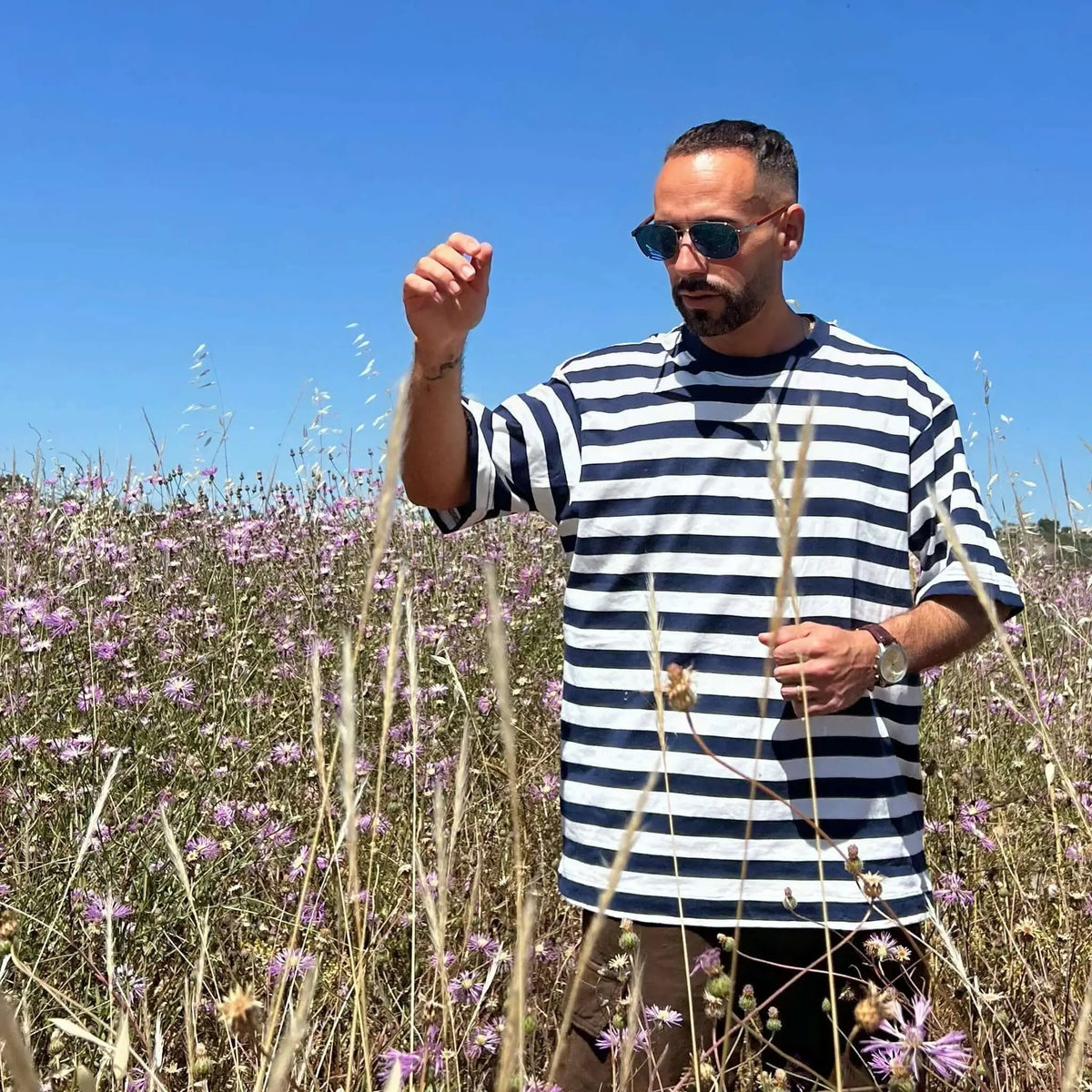 Hombre con gafas de sol de lentes azules y patillas de madera en un campo de flores, estilo casual y veraniego.
