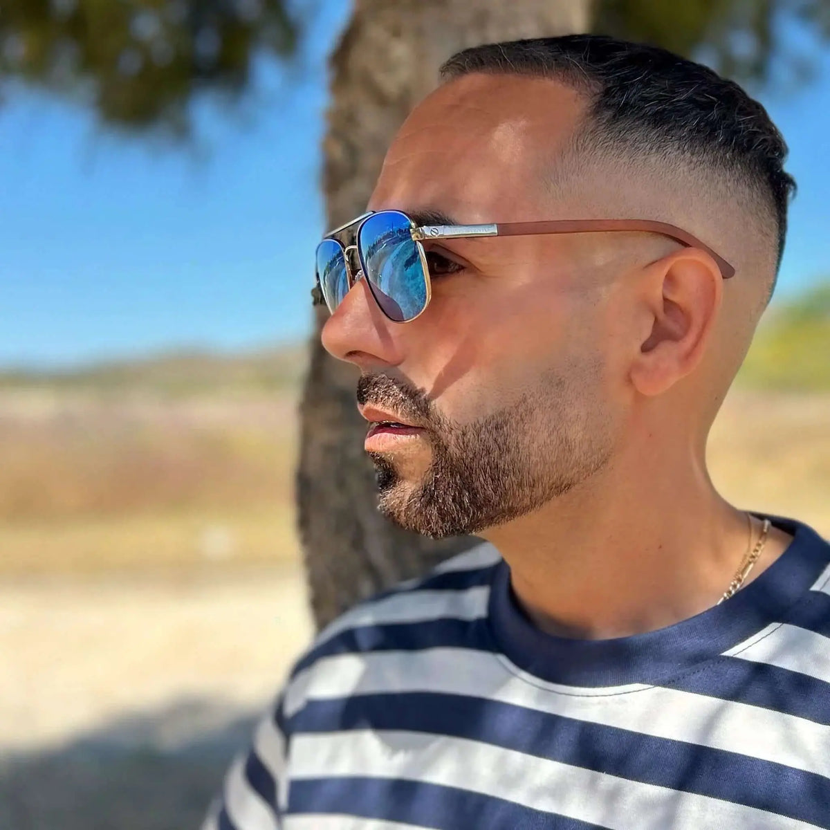 Hombre con gafas de sol de montura metálica, lentes azules espejadas y patillas de madera, en exteriores soleados.