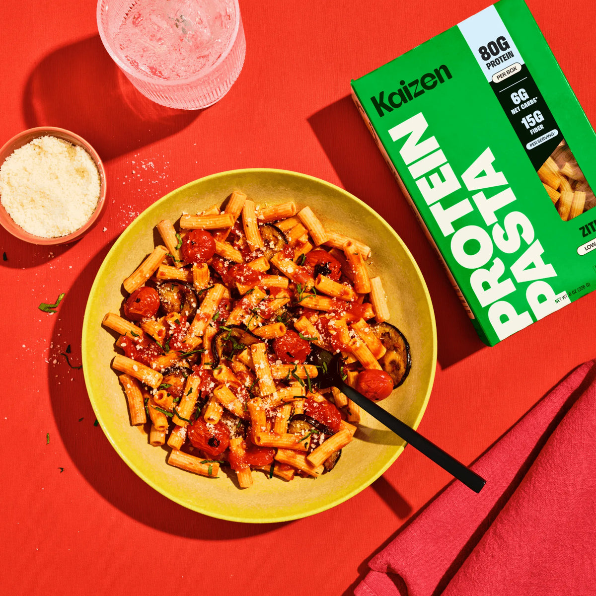 A bowl of ziti with tomato sauce, eggplant, and herbs next to a Kaizen Protein Pasta box on a bright red background.