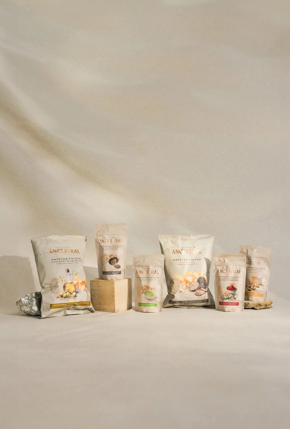Six gourmet snack bags arranged on neutral blocks and stone, featuring clean beige packaging with elegant labels.