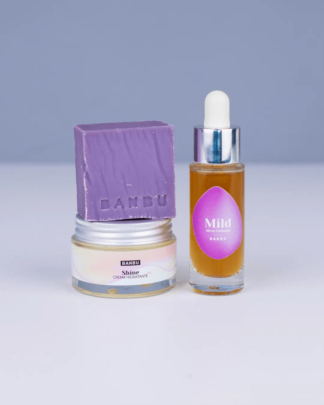 Pack facial Banbu: jabón morado, crema Shine en tarro de vidrio y sérum Mild con gotero sobre fondo liso azulado.