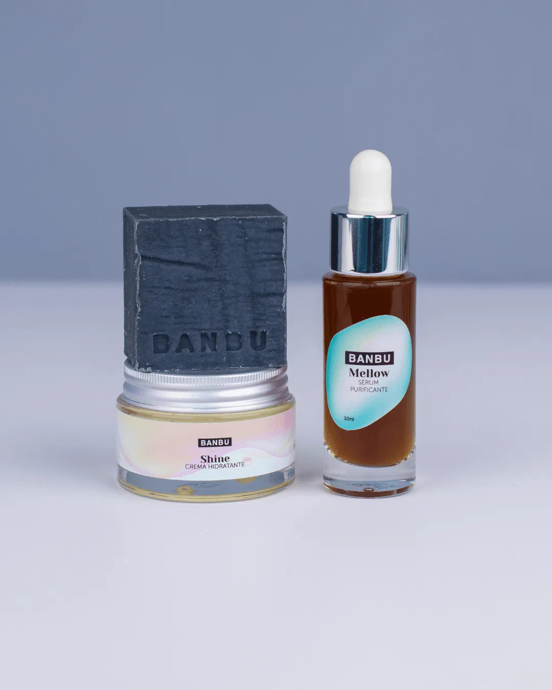 Pack Banbu: jabón gris oscuro, tarro de crema Shine y sérum Mellow en frasco de vidrio con cuentagotas sobre fondo gris.