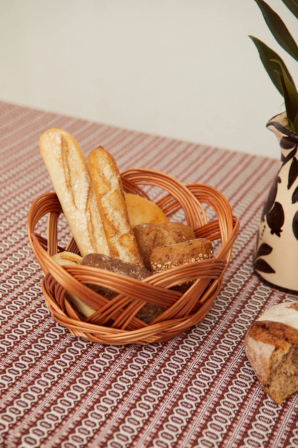 Round, open-weave natural wicker bread basket filled with assorted bread loaves on a textured patterned tablecloth.
