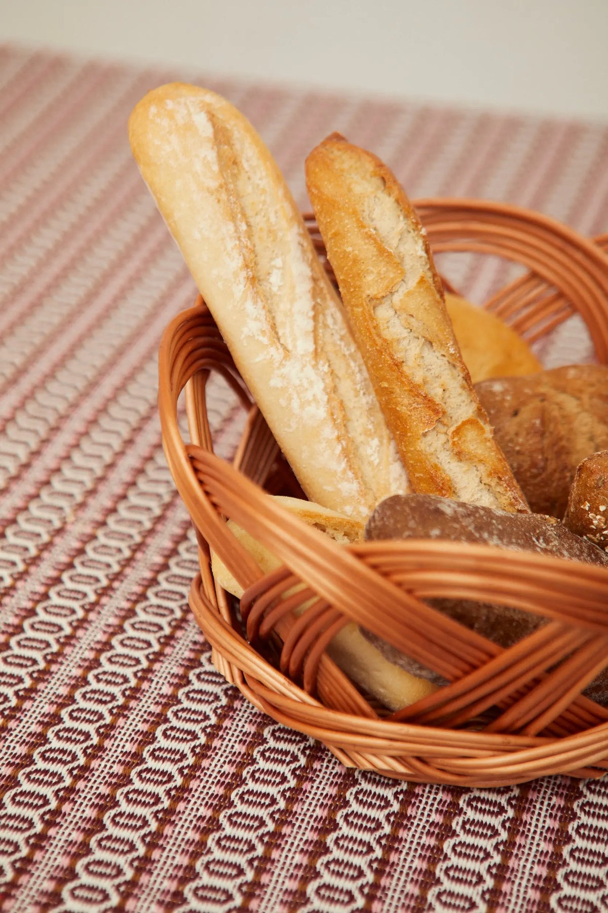 Round, open-weave natural wicker bread basket filled with various artisanal breads on a patterned fabric background.