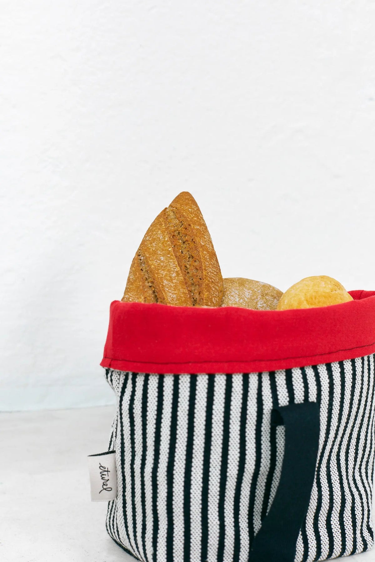 Panera de tela a rayas negras y blancas, con borde superior rojo y asa lateral, llena de diversos tipos de pan.