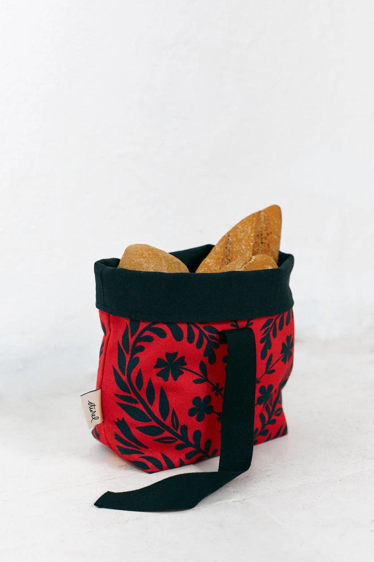 Panera de tela roja con estampado floral negro, estilo folk, con borde superior y asa en color negro, llena de pan.