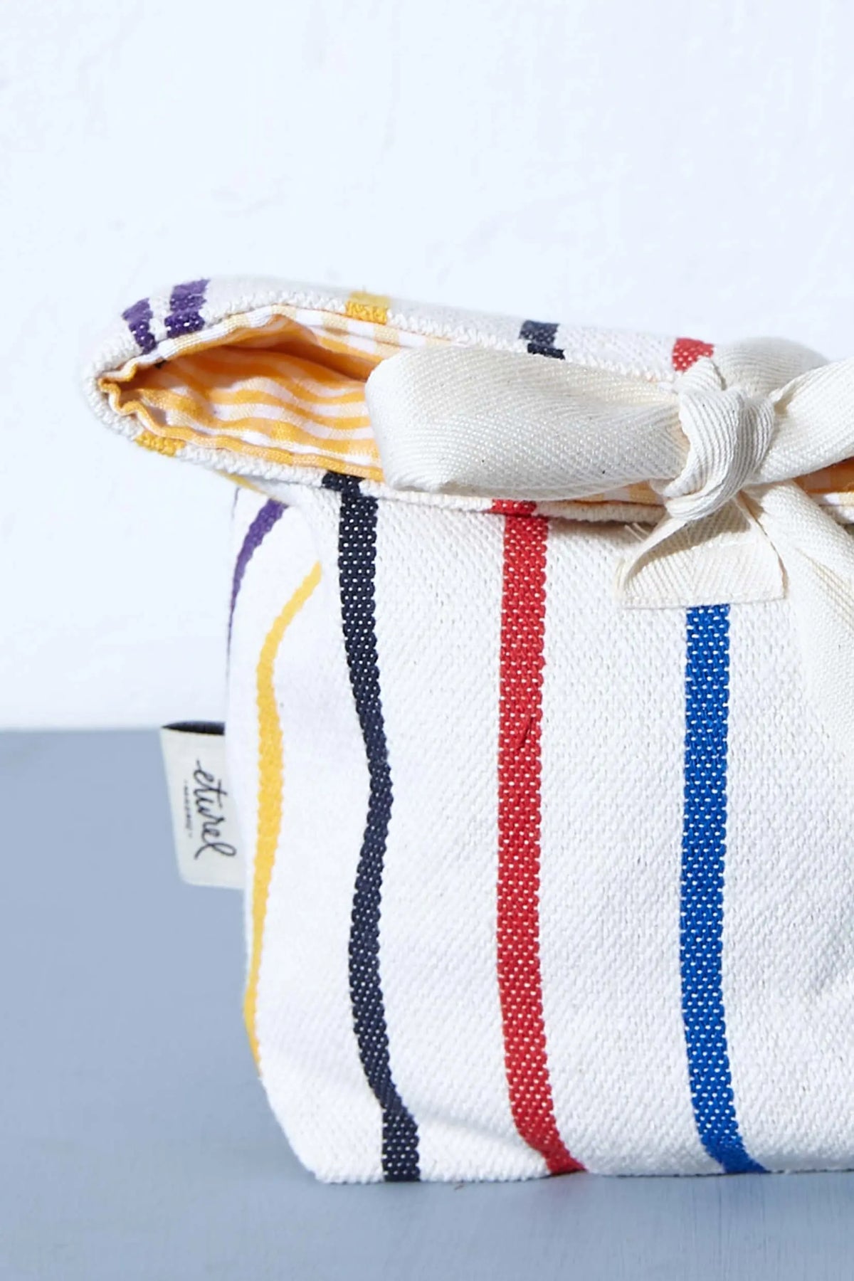Panera de tela blanca con rayas verticales de colores, cierre con lazo y forro interior a cuadros amarillos.