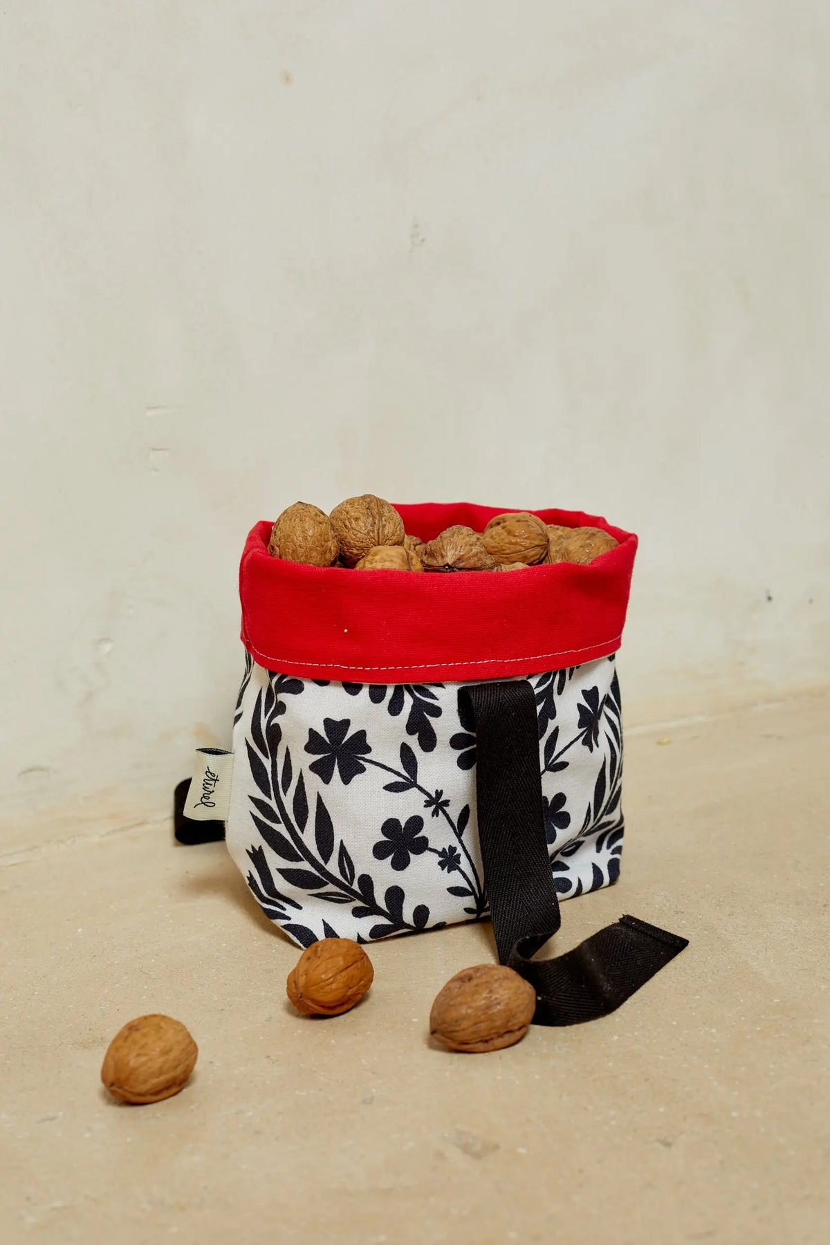 Panera de tela blanca con estampado floral negro, borde superior rojo y nueces en su interior.