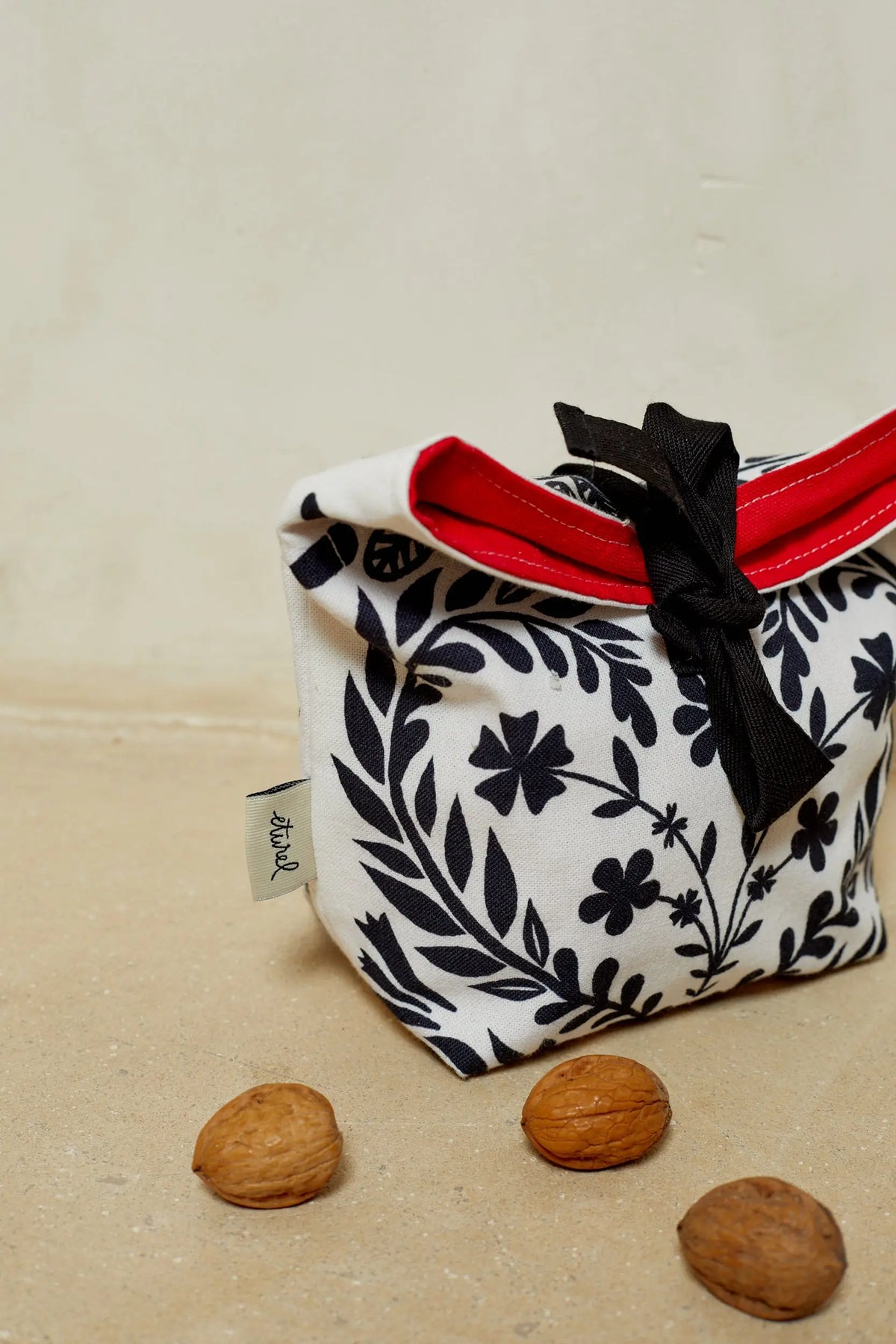 Panera de tela blanca con estampado floral negro, borde interior rojo y cierre con lazo negro sobre fondo neutro.