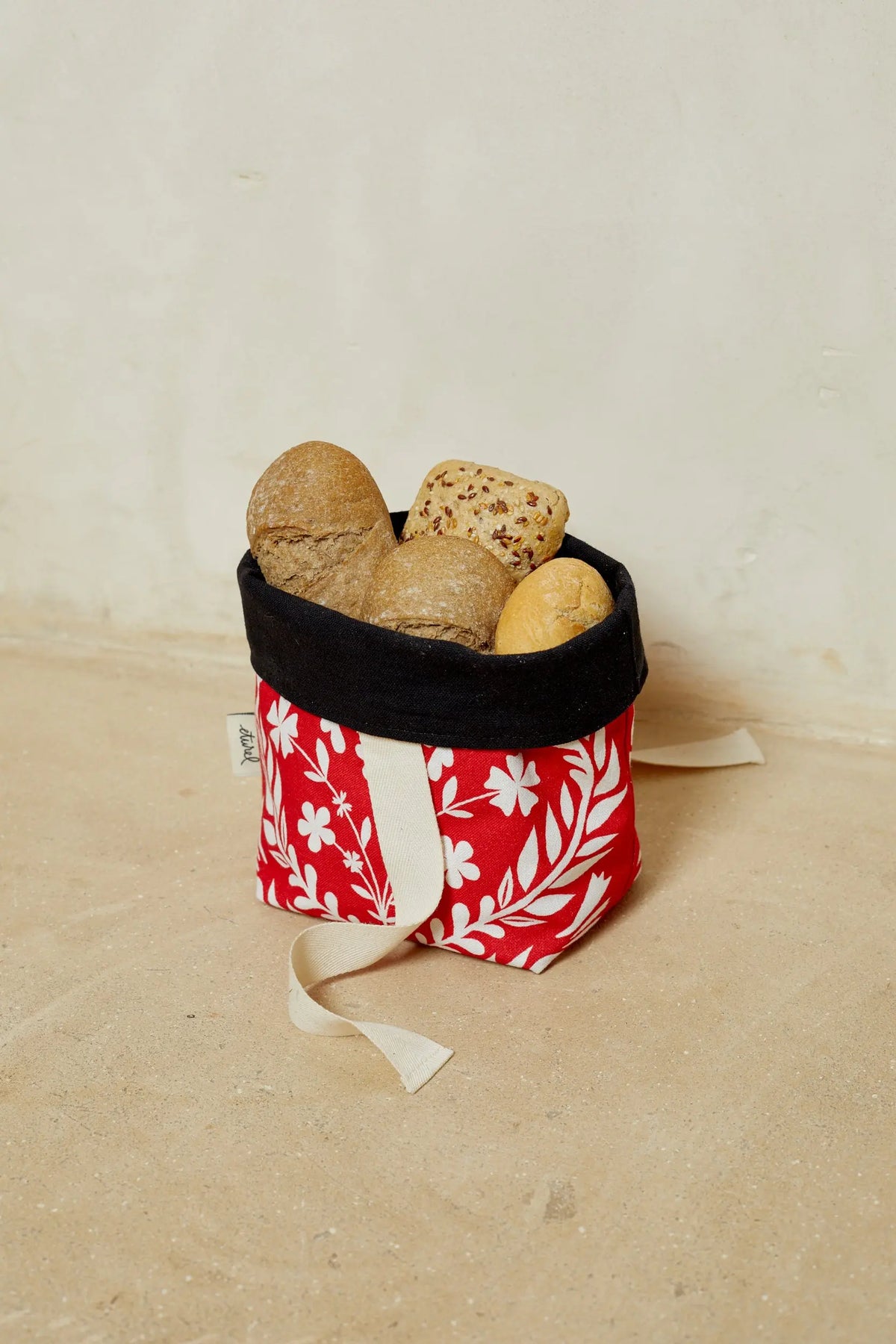 Small red fabric bread basket with white floral print, black interior lining, and cream-colored straps filled with bread.