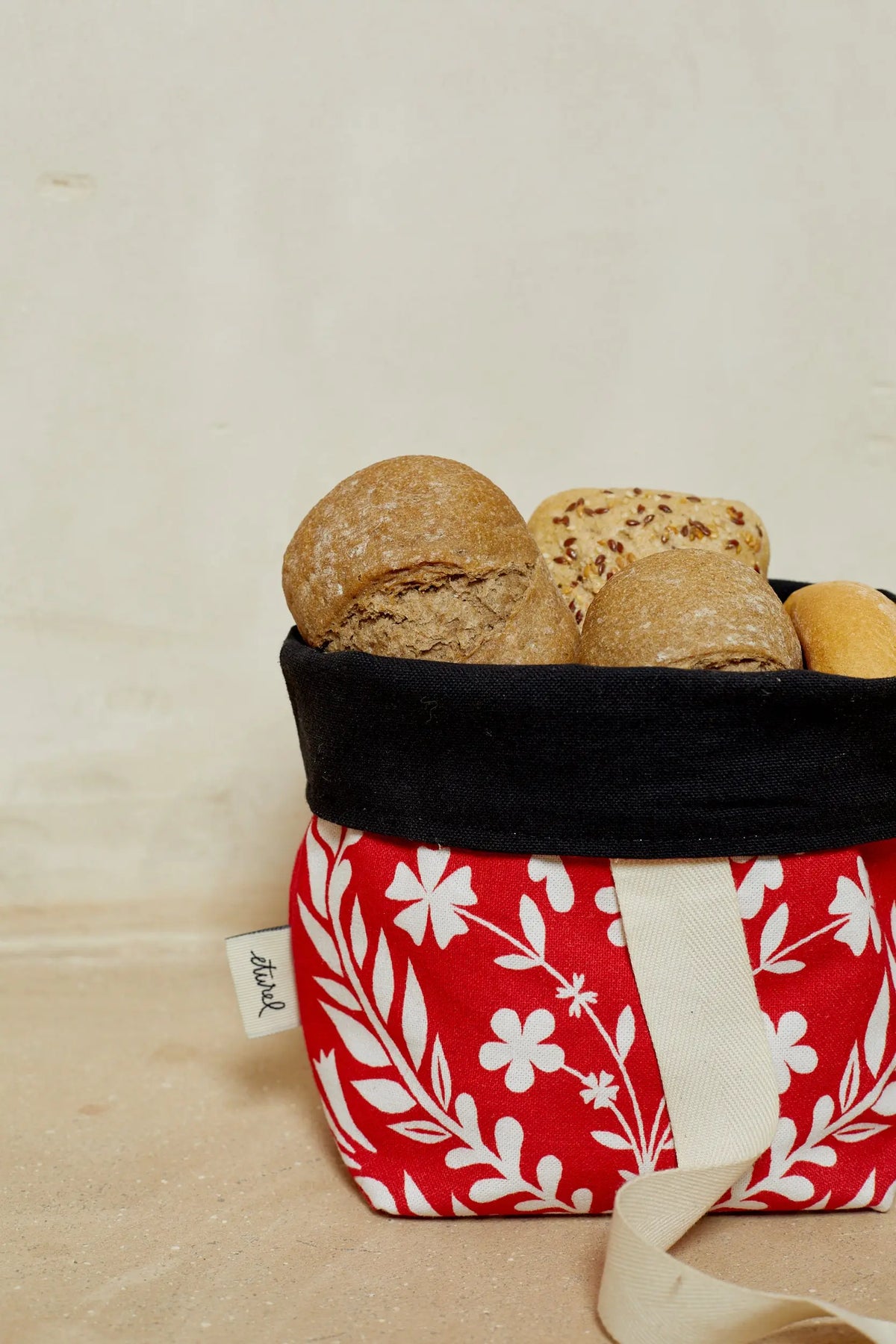 Panera de tela roja con estampado floral blanco, borde negro y asas color crema, llena de diferentes tipos de pan.