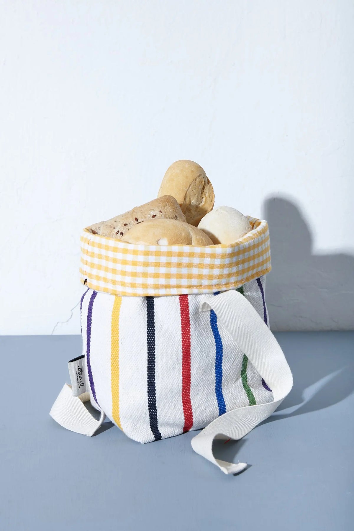 Panera de tela con rayas coloridas y borde interior de cuadros amarillos, llena de piezas de pan sobre fondo azul.