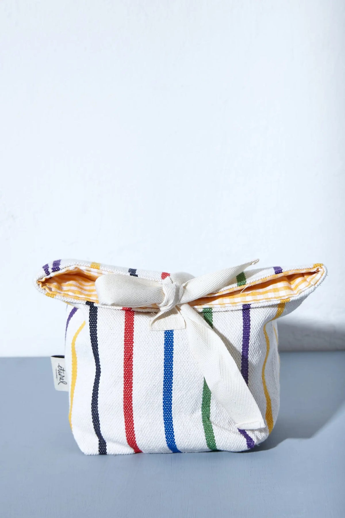 Panera de tela blanca con rayas verticales de colores y lazo de cierre, interior con forro de cuadros amarillo.