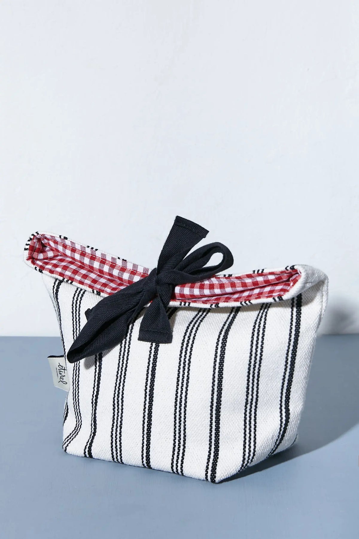 Panera de loneta con rayas negras, interior de cuadros vichy rojos y lazo negro decorativo sobre fondo gris.