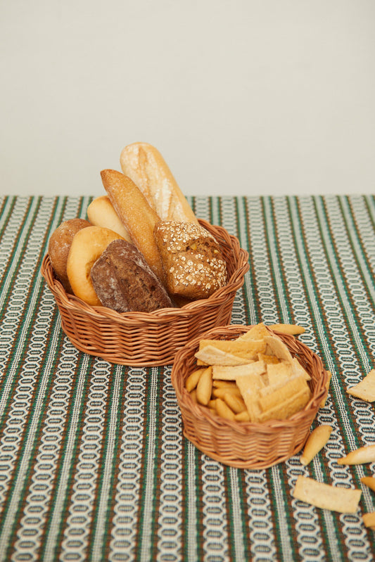 Two round, woven natural wicker baskets filled with assorted breads and crackers on a striped green patterned cloth.