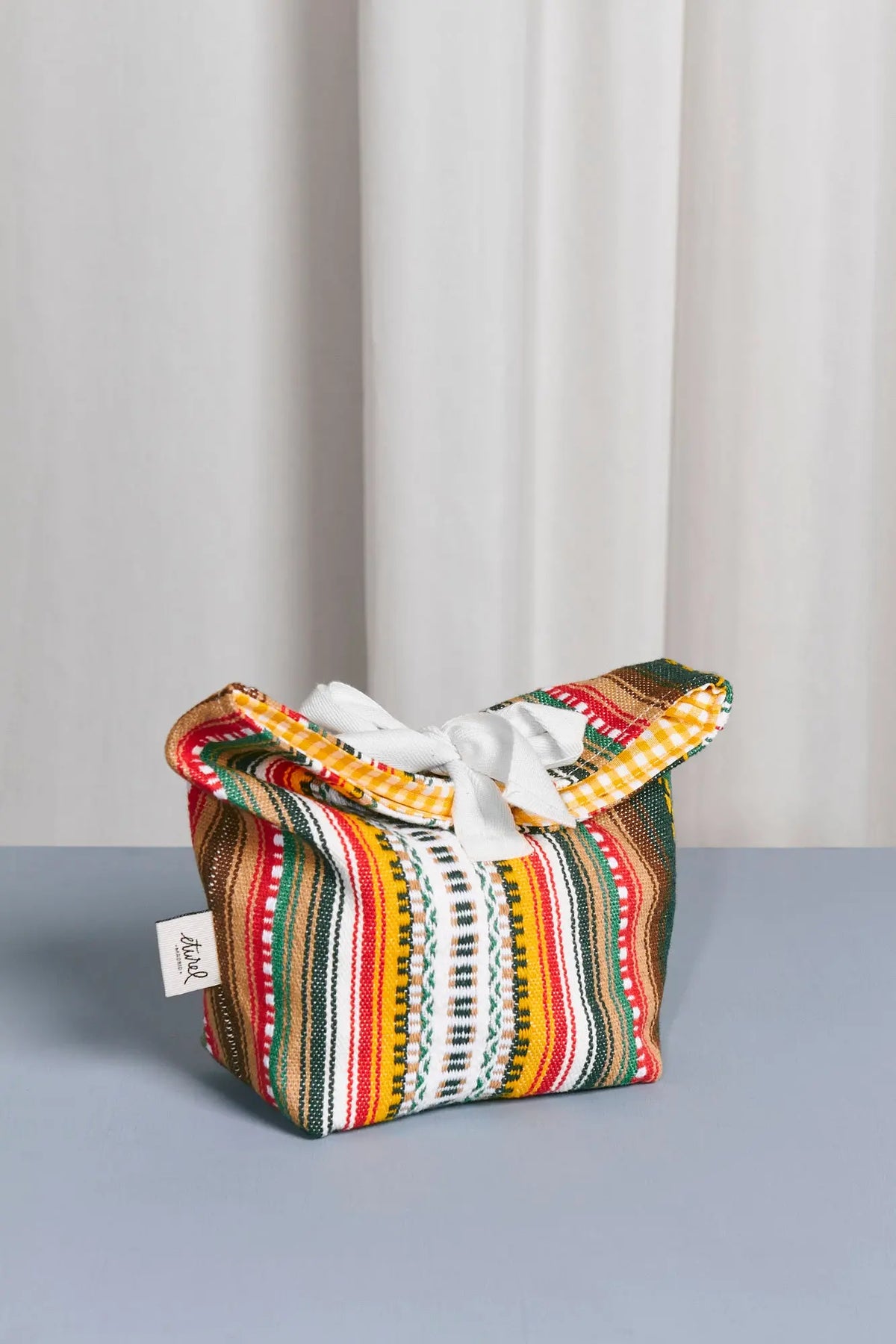 Panera de tela con estampado tradicional multicolor a rayas, estilo rústico, con cierre de lazo blanco superior.