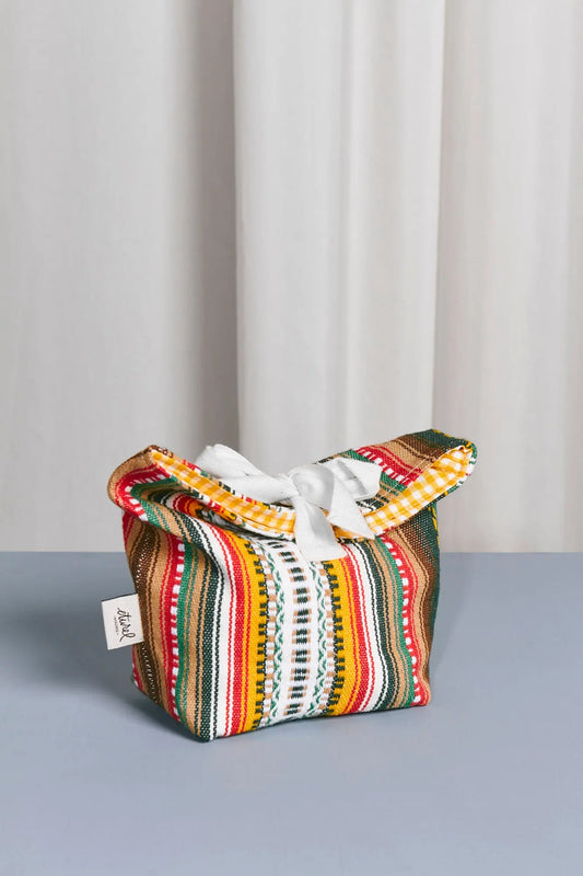 Panera de tela con estampado tradicional multicolor a rayas, estilo rústico, con cierre de lazo blanco superior.