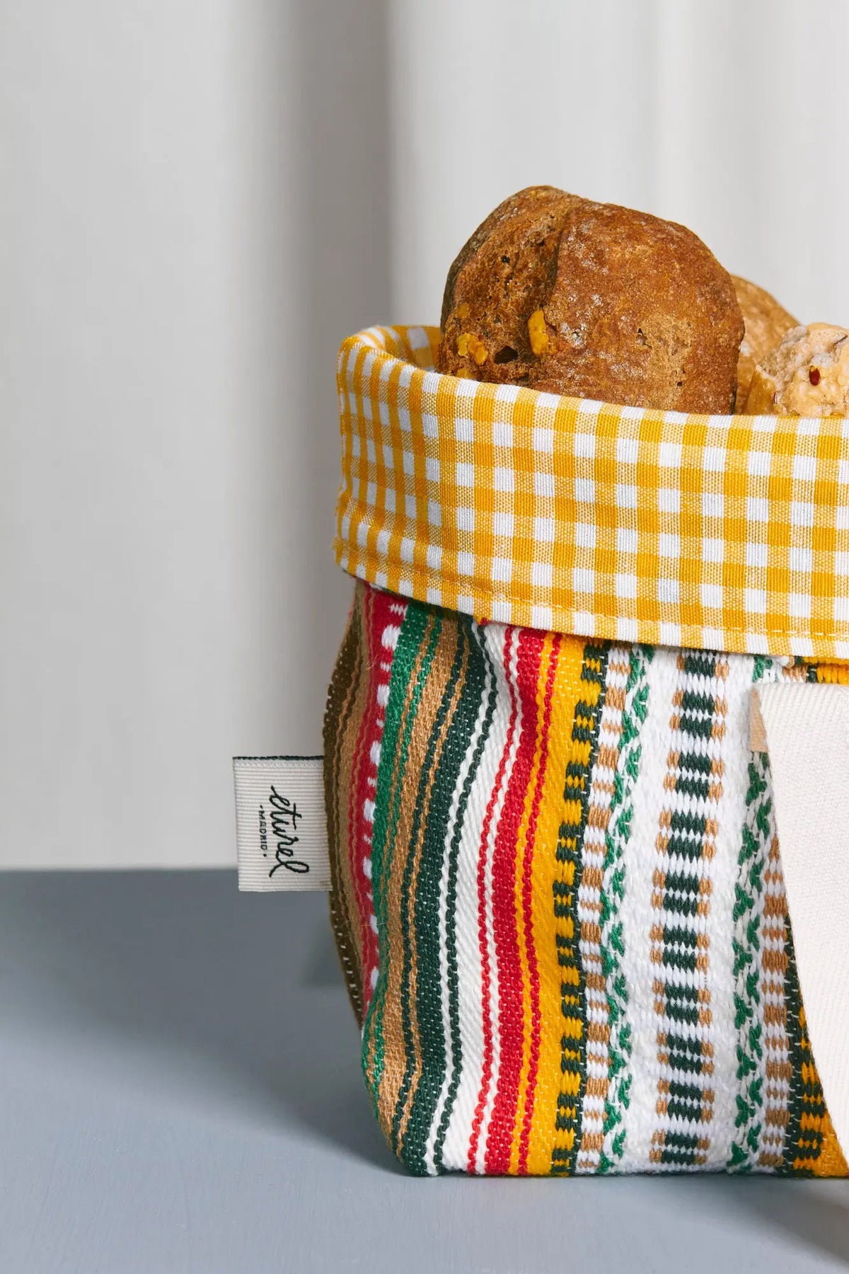 Panera de tela con estampado de rayas coloridas y borde superior de cuadros vichy amarillos, rellena con pan artesano.