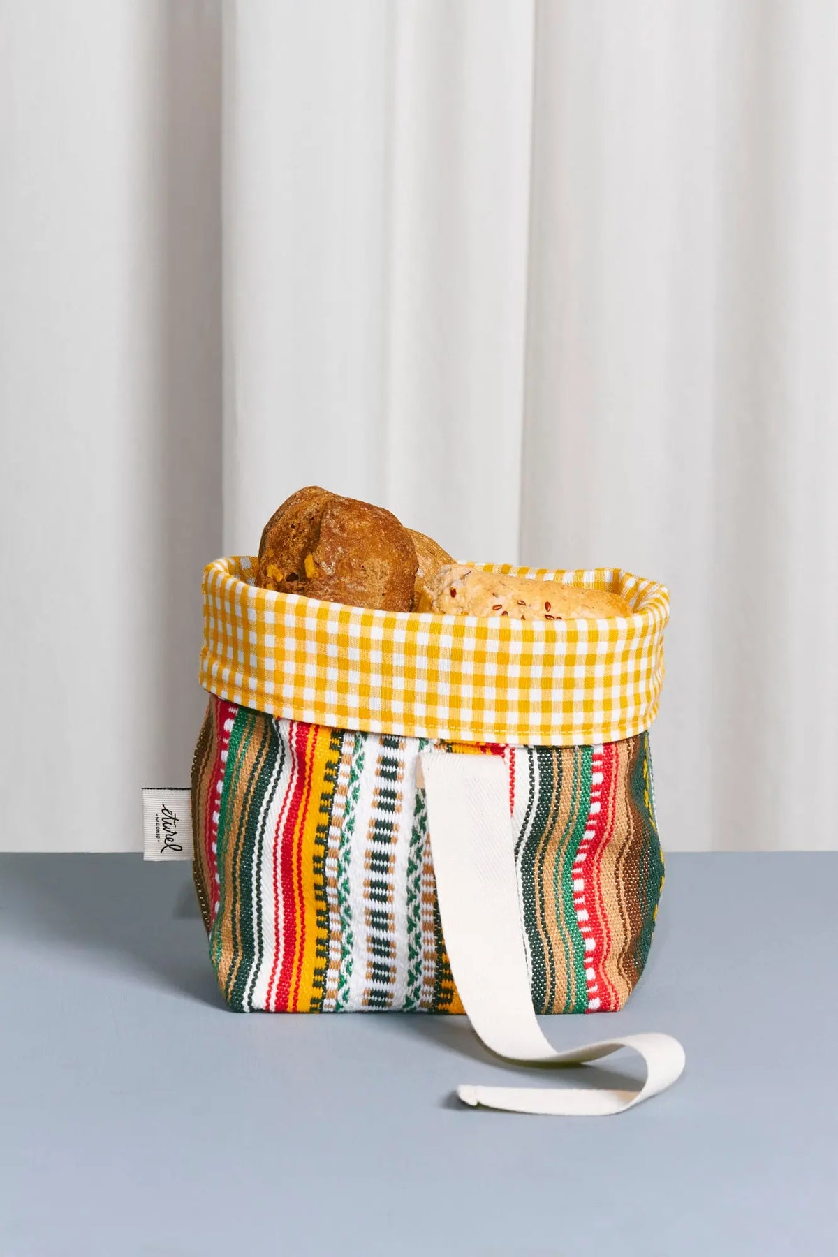 Panera pequeña de tela con estampado alpujarreño multicolor y borde superior de cuadros amarillos, llena de pan.