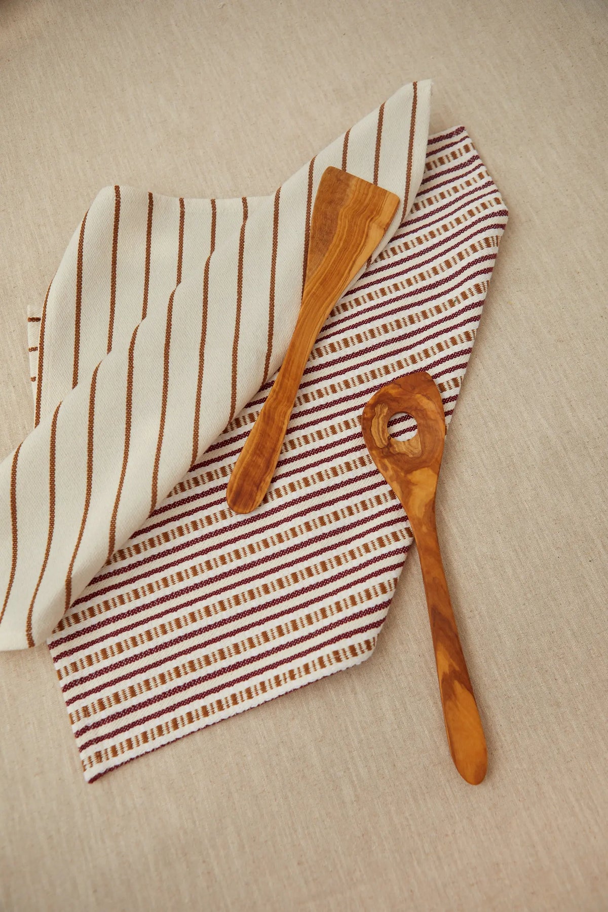 Set de dos paños de cocina de algodón con rayas granates y beige, estilo rústico, acompañados de utensilios de madera.