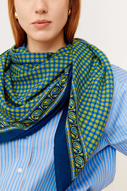 Blue and green patterned silk scarf styled on a model, paired with a light blue striped shirt.
