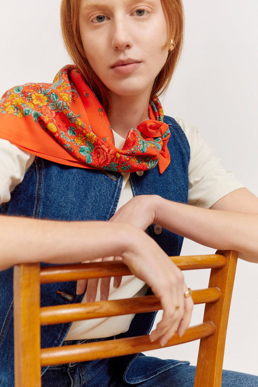 Red square scarf with a vibrant floral pattern in yellow and green, worn around the neck by a woman in a denim vest.